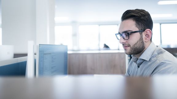dunkelhaariger Mitarbeiter mit Brille in Büro an Schreibtisch vor PC Bildschirm Hintergrund Fensterfront hell unscharf 