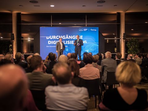 Publikum sitzt vor einer Bühne, darauf die Geschäftsleitung Jürgen Häring unterhaltet sich mit einem dunkelhaarigem Mann